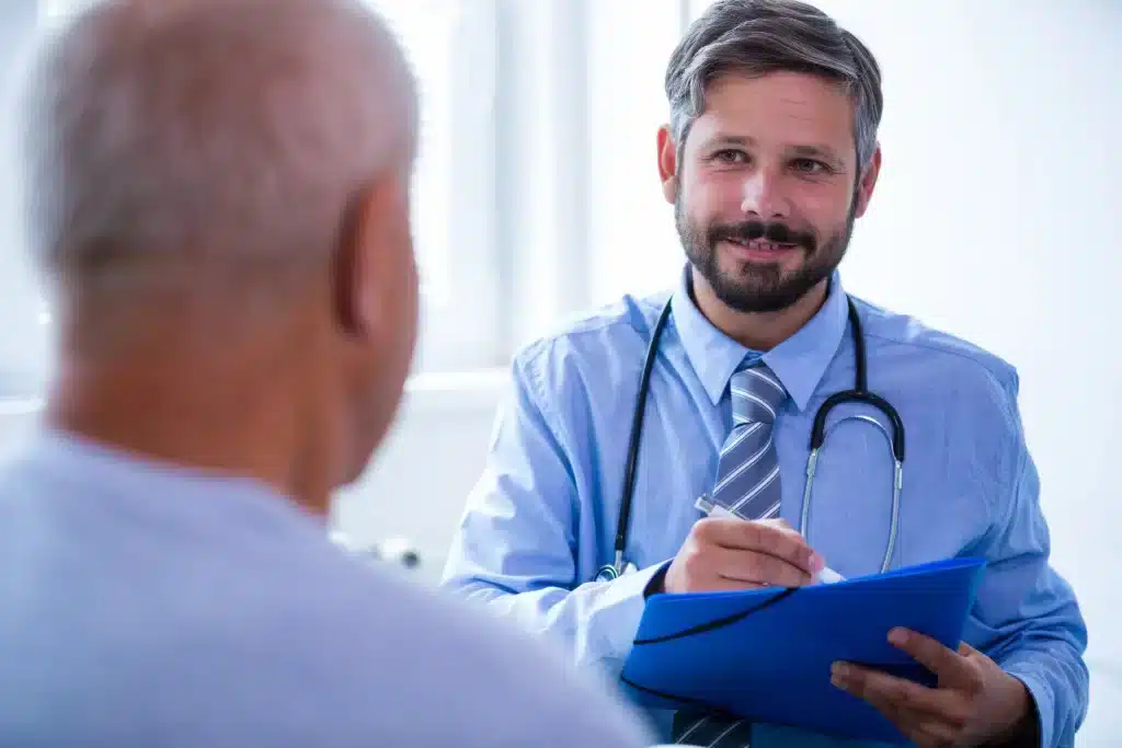 consulta-revision-urologica-merida-en-consultorio Médico urológico en Mérida escuchando atentamente a su paciente adulto mayor durante una consulta de revisión urológica en consultorio privado.